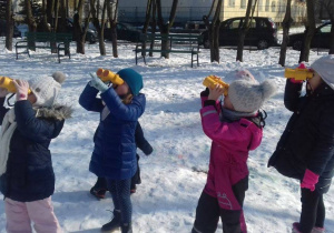 Ogród przedszkolny w zimowej szacie, dzieci z lornetkami prowadzą obserwacje przyrodnicze.